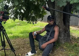 Un jeune homme assis sous un arbre. On aperçoit à gauche une caméra et à droite un micro.
