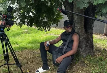 Un jeune homme assis sous un arbre. On aperçoit à gauche une caméra et à droite un micro.