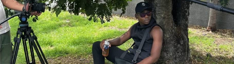 Un jeune homme assis sous un arbre. On aperçoit à gauche une caméra et à droite un micro.